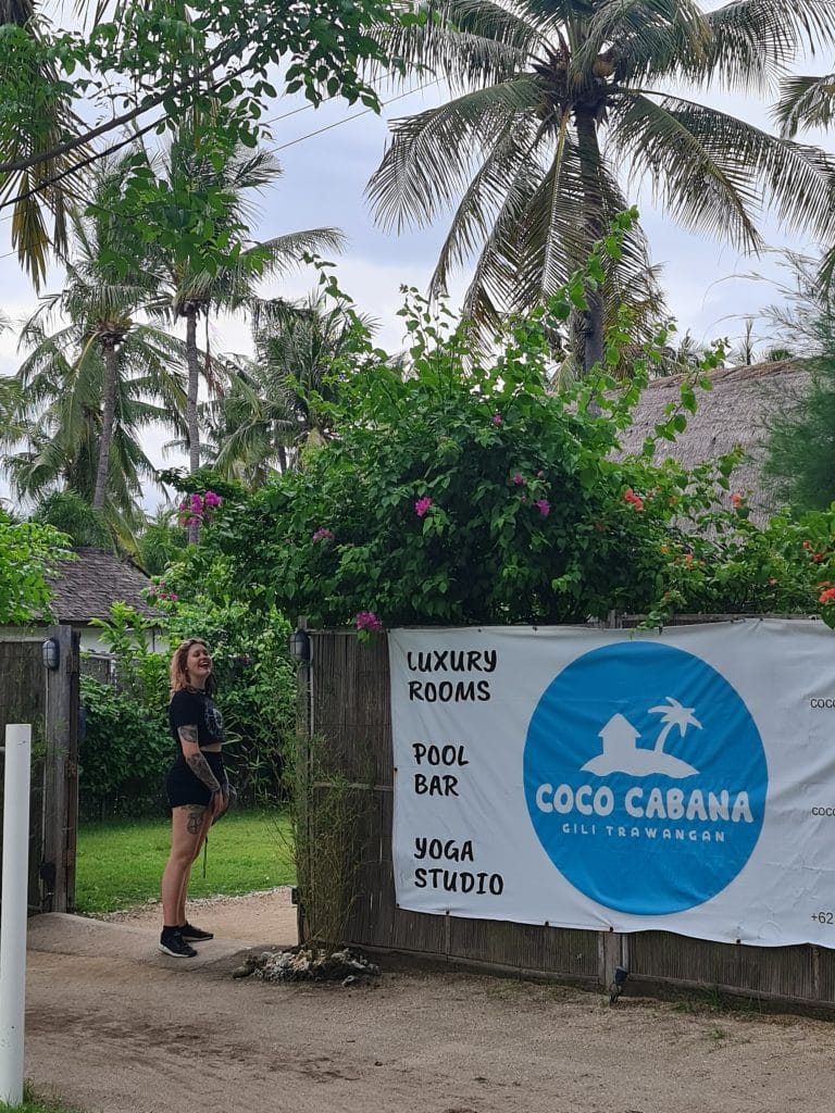 Sover Travel - A person stands near a fence at the entrance of Coco Cabana on Gili Trawangan, where elegant architecture highlights luxury rooms, a pool bar, and a yoga studio. Palm trees and tropical greenery frame the sunlit streets leading to this serene escape.