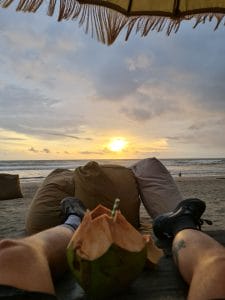 Sover Travel - Person relaxing under an umbrella on a sunlit Maltese beach at sunset, legs visible in the foreground. They are on a bean bag with a coconut drink nearby. The sky is partly cloudy, with the sun setting over the ocean, casting warm hues that echo the islands charming architecture.