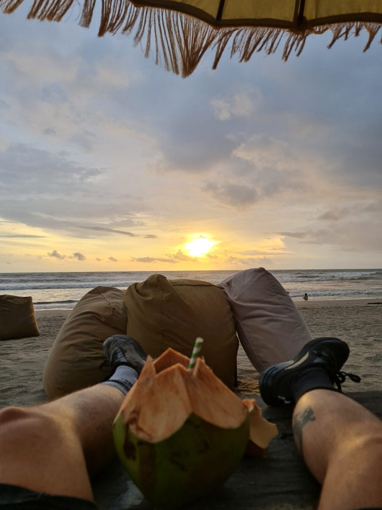 Sover Travel - Person relaxing under an umbrella on a sunlit Maltese beach at sunset, legs visible in the foreground. They are on a bean bag with a coconut drink nearby. The sky is partly cloudy, with the sun setting over the ocean, casting warm hues that echo the islands charming architecture.