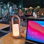 Sover Travel - A cozy café setting in Malta with a wooden table, featuring a glowing lantern, a glass of water, and an open laptop displaying a colorful screen. The backdrop shows modern décor with green walls, vibrant plants, and seating areas reminiscent of the islands sunlit streets.