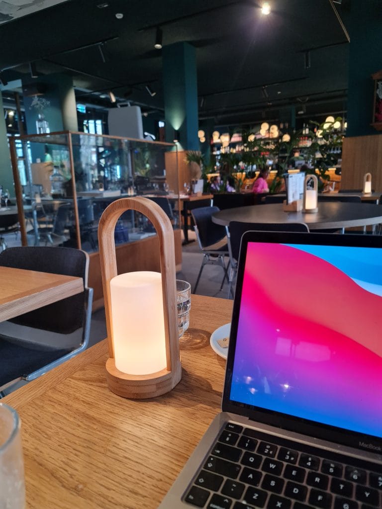 Sover Travel - A cozy café setting in Malta with a wooden table, featuring a glowing lantern, a glass of water, and an open laptop displaying a colorful screen. The backdrop shows modern décor with green walls, vibrant plants, and seating areas reminiscent of the islands sunlit streets.