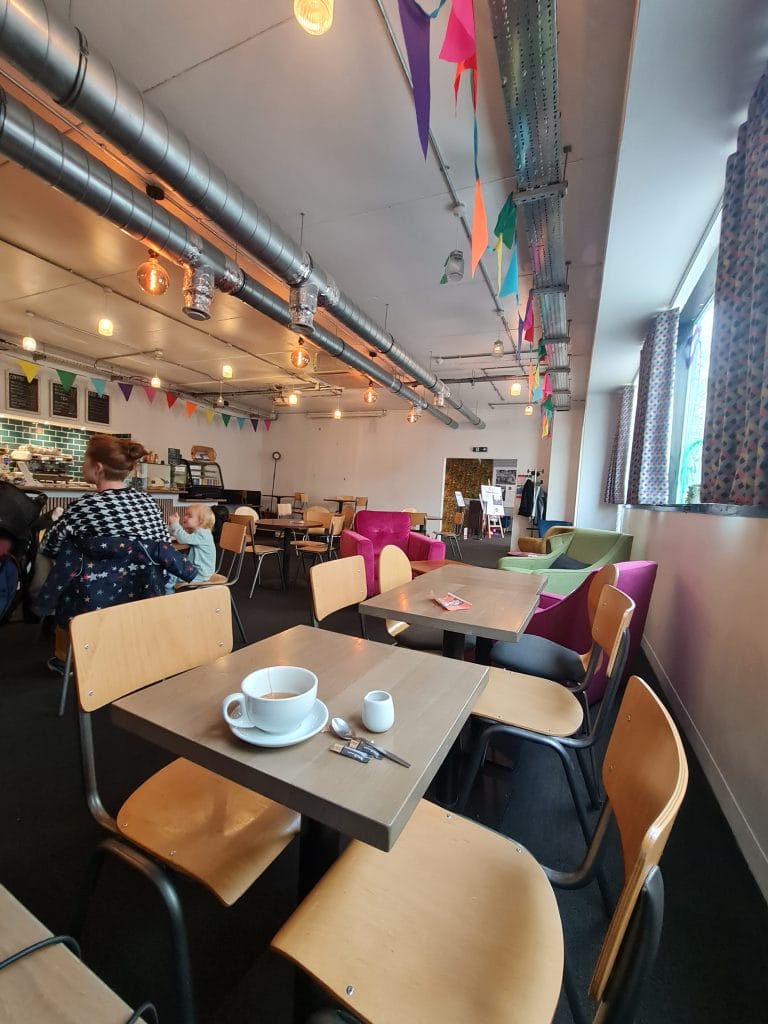 Sover Travel - A cozy café with modern decor is nestled along the sunlit streets of Malta, featuring colorful triangular flags hanging from the ceiling. Wooden tables and chairs are set up, with a cup and saucer on one table. Patrons sit at tables, and the counter is visible in the background.