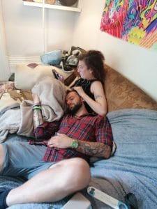 Sover Travel - A man and a woman are lounging on a cozy sofa. The woman is sitting up, holding a phone, while the man rests his head on her lap, sleeping. A colorful abstract painting is on the wall behind them. The scene is relaxed and intimate.