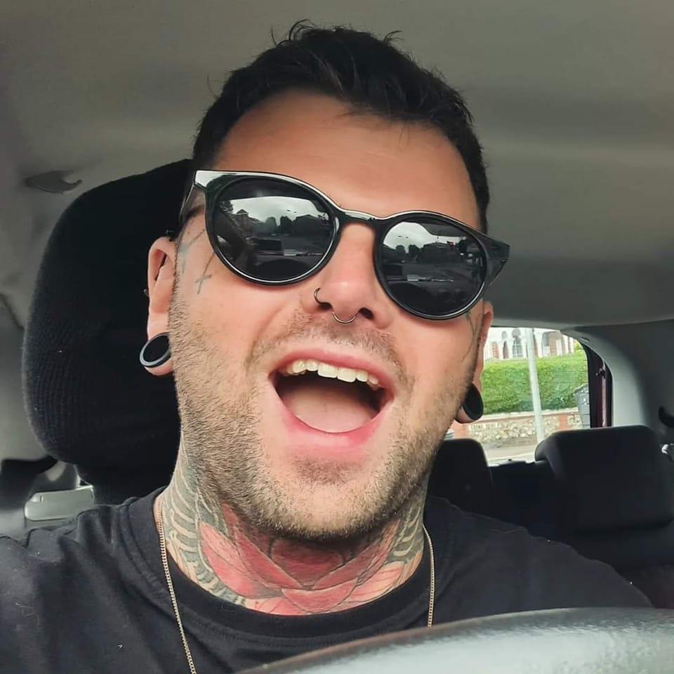 Sover Travel - A person with dark hair, sunglasses, and a nose piercing is sitting in a car, smiling widely. They have tattoos on their neck and earrings in both ears. The car window shows a blurry background with greenery and a building.
