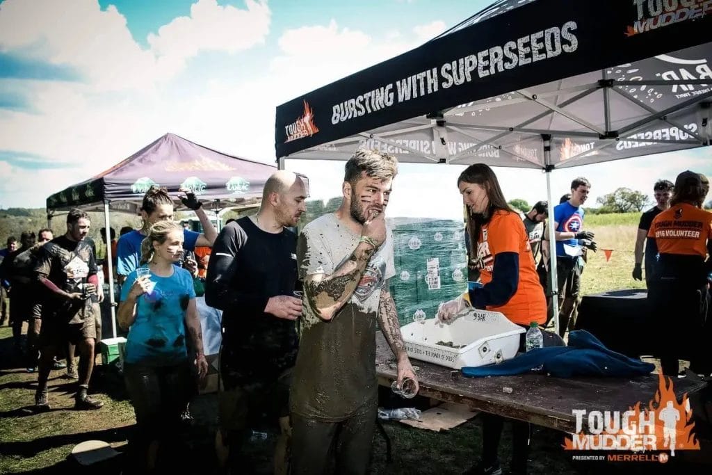 Sover Travel - Participants covered in mud at a Tough Mudder event sport vibrant neon gear as they grab snacks and drinks from a refreshment station. They appear tired but cheerful under a tent with branding, set against Camdens iconic backdrop and a clear blue sky.