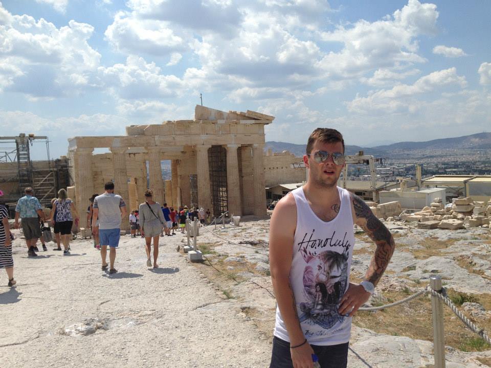 Sover Travel - A man in a tank top stands near ancient ruins, including the Parthenon, under a partly cloudy sky. Tourists explore the historical site in the background.