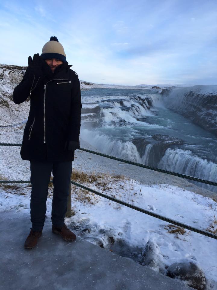 Sover Travel - Person in winter clothing waving on an icy path near a large waterfall with cascading water. The background shows a snowy landscape under a cloudy sky.