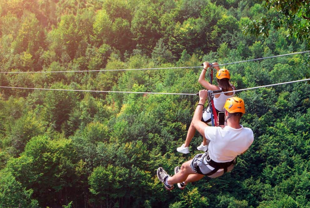 Sover Travel - Two people wearing orange helmets and harnesses zip line through a lush green forest. The bright sunlight filters through the trees, casting a warm glow on this adventurous scene. It’s one of the best activities for those seeking alcohol-free travel and thrilling experiences.