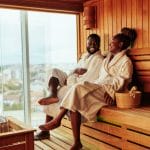 Sover Travel - Two people in white robes sit on a wooden bench in a sauna, smiling and conversing about exciting destinations. A wooden bucket is beside them, and a large window reveals a city view beyond. This serene setting proves exploring without alcohol can be equally delightful.