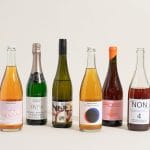 Sover Travel - A lineup of eight non-alcoholic beverage bottles, featuring an array of sparkling wines and drinks, is displayed on a neutral background. Each bottle boasts local flavors with distinct labels and designs, from transparent to colorful, celebrating diversity in branding for the sober traveller.