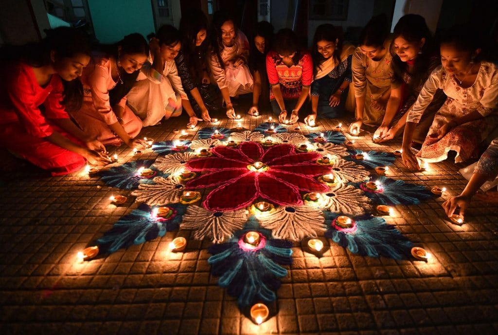 **Alt Text:** A group of women lighting candles around a colorful rangoli design during a festive celebration. **Caption:** Celebrate the joy of community and mindfulness as these women create a beautiful rangoli, enhancing their festive spirit. **Image Description:** Women gather to light candles around a vibrant rangoli, symbolizing unity and celebration.