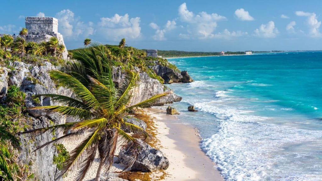 Sover Travel - Experience the scenic view of Tulums beach, where turquoise waters meet white waves along a rocky shoreline. Set against ancient Mayan ruins and swaying palm trees, this destination is perfect for your Sober Travel Wishlist under a partly cloudy blue sky.