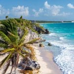 Sover Travel - Experience the scenic view of Tulums beach, where turquoise waters meet white waves along a rocky shoreline. Set against ancient Mayan ruins and swaying palm trees, this destination is perfect for your Sober Travel Wishlist under a partly cloudy blue sky.