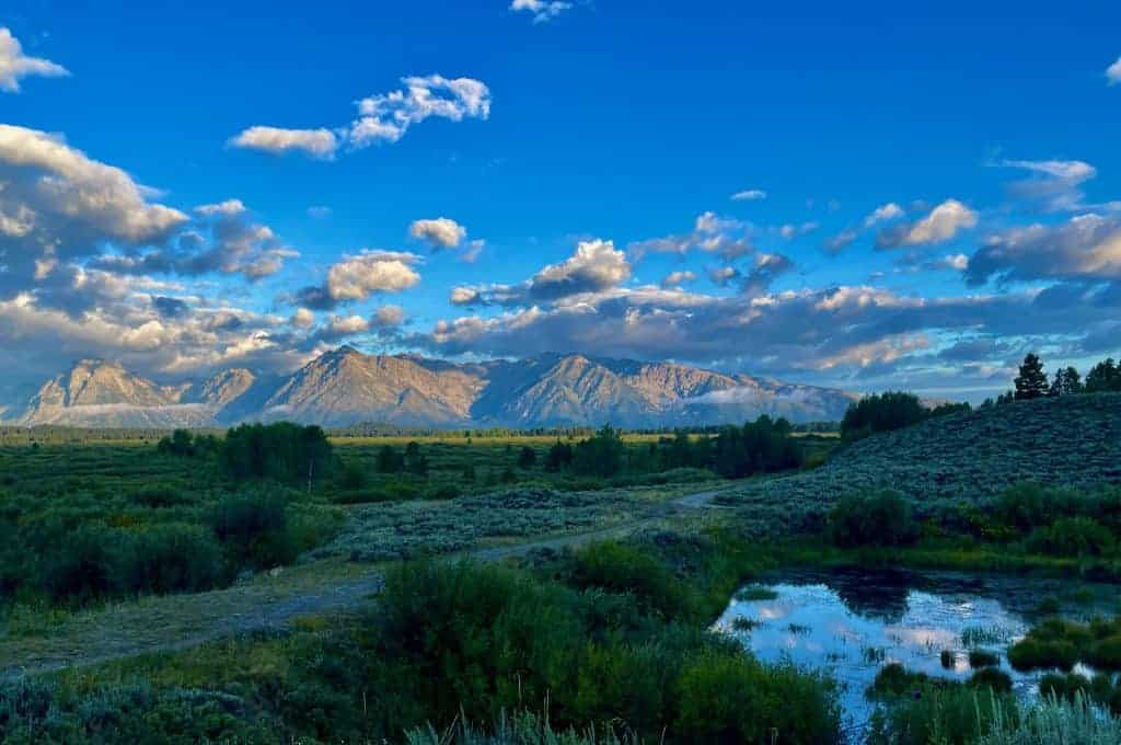 Sover Travel - Bask in the serenity of a vast landscape with lush greenery in the foreground and a reflective pond. Majestic mountains, one of the best destinations for mindful travel, are illuminated by sunlight under a vibrant blue sky adorned with scattered clouds.