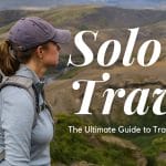 Sover Travel - A person with a backpack gazes at a vast, mountainous landscape. The words Solo Travel: The Ultimate Guide to Traveling Alone appear on the image. With journey confidence and wearing a cap and outdoor gear, they embrace the adventure ahead.