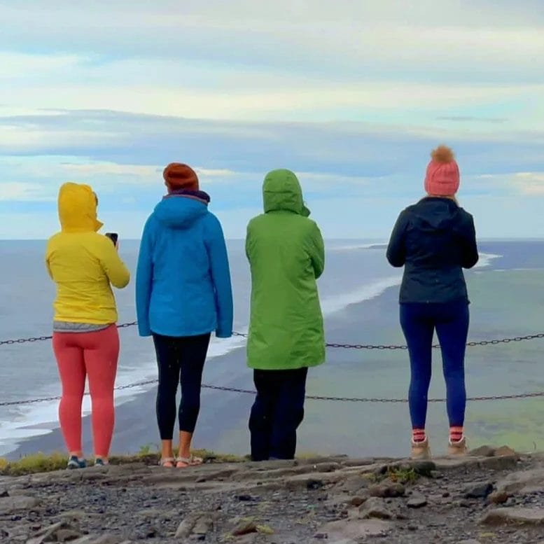 Sover Travel - Four people in colorful jackets stand on a cliff, embracing a moment of mindful travel as they overlook an ocean view. The sky is partly cloudy, and the sea stretches out in the distance. A safety chain fence runs along the edge.