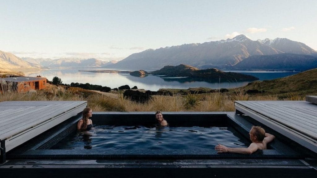 Sover Travel - Three people unwind in a Healing Space, a hot tub overlooking a scenic lake and mountain range. The sky is clear, and the sun sets behind the distant peaks. Two people are seated on the left, one reclines on the right, all reveling in this tranquil, immersive Sober Retreat experience.