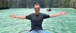 Sover Travel - A man with sunglasses and a broad smile sits at the front of a boat, arms outstretched in pure joy, exploring the world. Hes surrounded by turquoise waters and lush, rocky cliffs, while several small boats dot the background—a perfect moment captured for his sober travel blog.