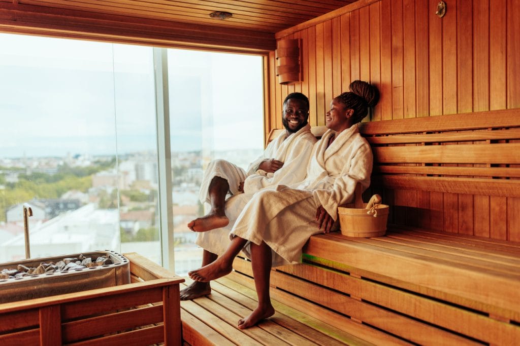 Sover Travel - Two people in white robes relax and smile while sitting on wooden benches inside a sauna at an alcohol-free destination. A basket with sauna items is nearby, complementing this enriching journey. A large window offers a view of a cityscape under a cloudy sky.