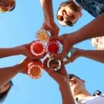 Sover Travel - A trend-setting group seen from below, smiling and holding colorful drinks in plastic cups against a clear blue sky. They are casually dressed, wearing sunglasses, and radiate a cheerful mood perfect for embracing the sober travel lifestyle.