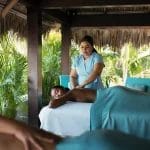 Sover Travel - In a tranquil, sober travel experience, a person luxuriates in a massage on a table amidst lush greenery. The therapist wears a light blue uniform, while another guest enjoys opulence without alcohol nearby.