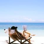 Sover Travel - A person in a sun hat relaxes on a lounge chair, engrossed in a book, enjoying an alcohol-free fun day on a sandy beach. The ocean is calm with clear blue water under a bright sky dotted with clouds—an ideal sober vacation escape.