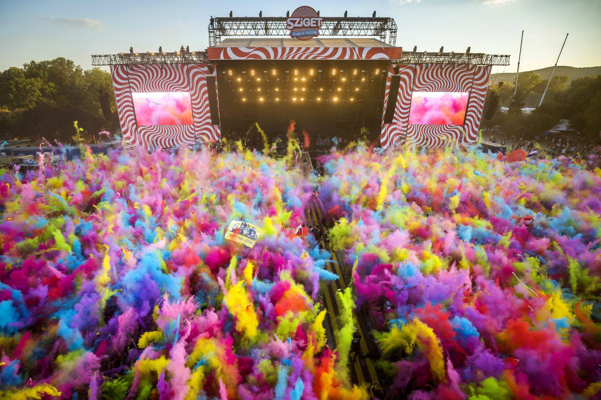 Sover Travel - A vibrant festival scene with colorful powder clouds filling the air. A large stage with Sziget signage is in the background, illuminated by lights. The crowd is lively and energetic, surrounded by a mix of bright pink, yellow, blue, and purple hues.
