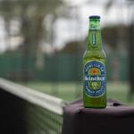 Sover Travel - A bottle of Heineken 0.0% non-alcoholic beer rests confidently on a tennis court net post, capturing TikTok-worthy vibes. In the blurred background, a green tennis court and fencing highlight the zero-proof refreshment with modern flair.